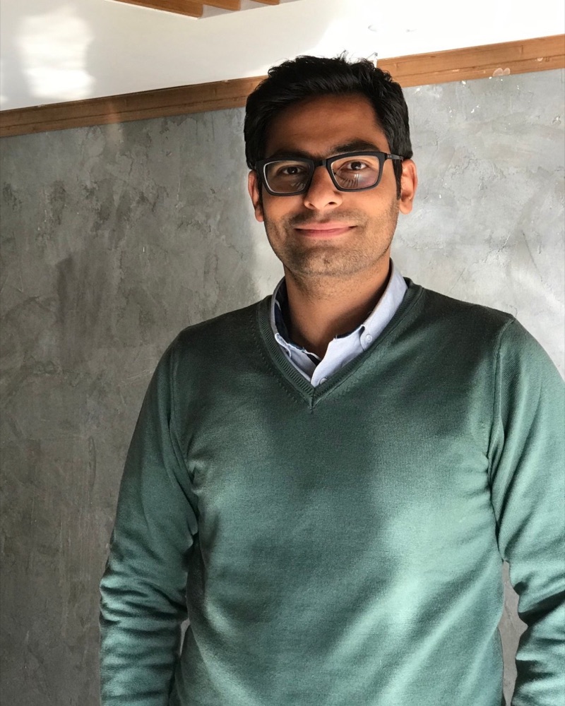 Sepehr Pandsheno, smiling, wearing glasses and a green sweater, standing in front of a grey plaster wall.